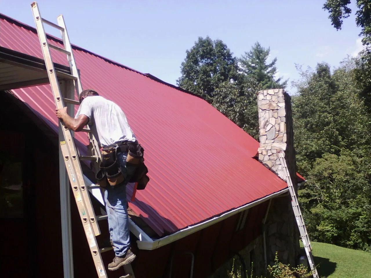 Roofer on a ladder beside a red metal roof and stone chimney — Soffit & Fascia Repair by Roofing Brothers in Big Lake